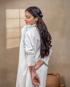 Model wearing a white laad`s midi cotton dress with pastel floral embroidery on the neckline and blue threadwork on the sleeves, featuring a ruffled hem and relaxed fit.