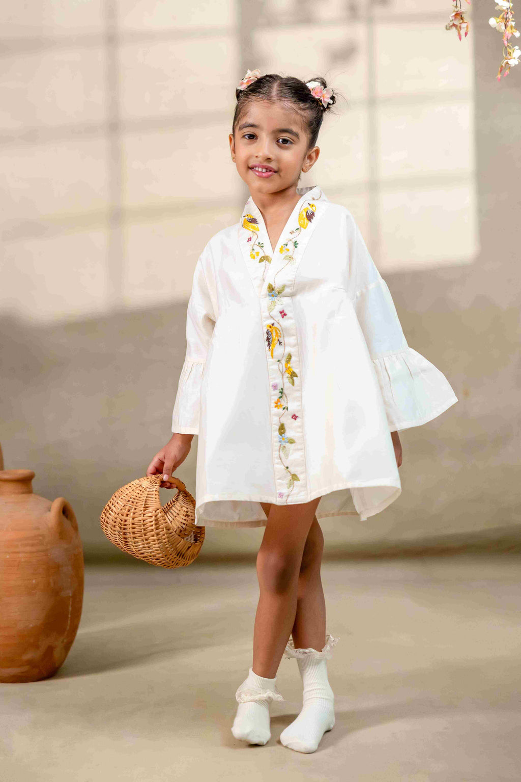Little girl in a white hand-embroidered A-line girls party dress with flared sleeves and colorful bird and floral detailing, holding a wicker basket.