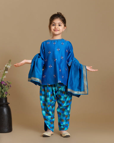 A young girl wearing  Laad`s  vibrant blue embroidered kurta with delicate floral motifs, paired with checkered blue and green patiala pants and a matching dupatta with gold detailing. 