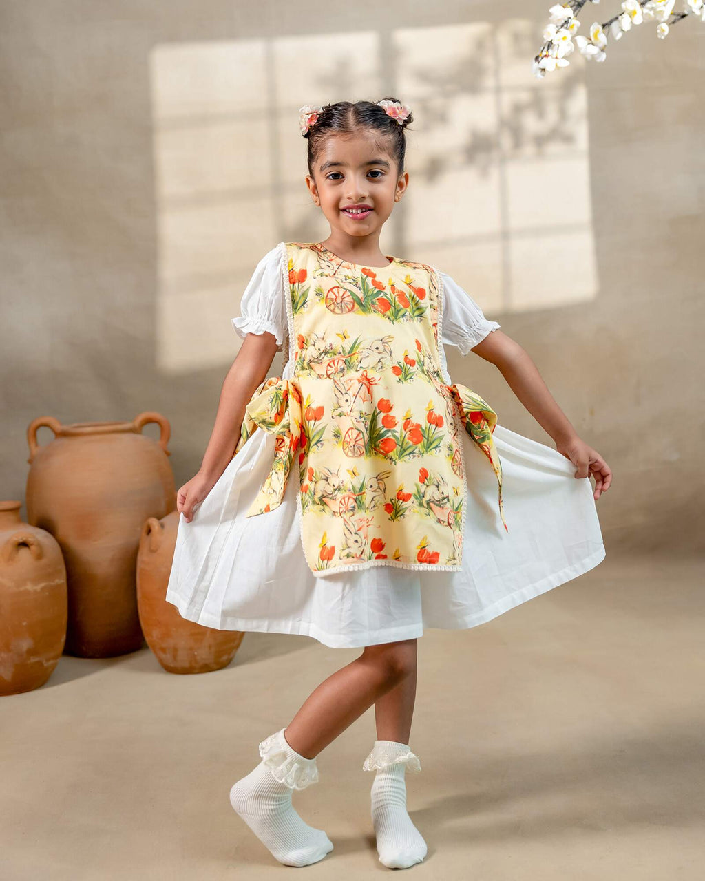 LAAD`S Girl in floral apron dress with a bunny and tulip print, layered over a white puff-sleeve dress. Features ruffled side panels and delicate lace trim. Soft, breathable fabric for comfort and charm.