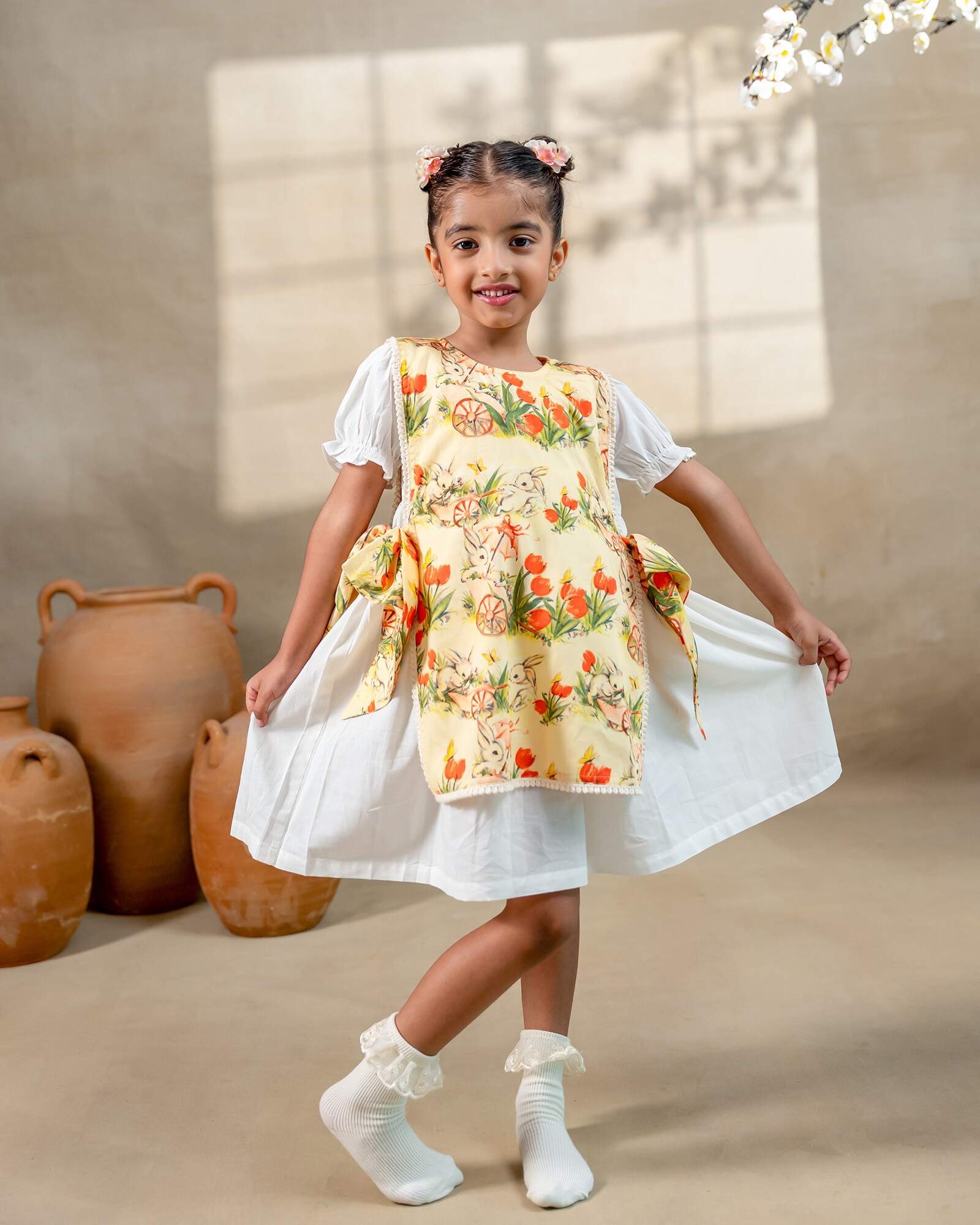 LAAD`S Girl in floral apron dress with a bunny and tulip print, layered over a white puff-sleeve dress. Features ruffled side panels and delicate lace trim. Soft, breathable fabric for comfort and charm.