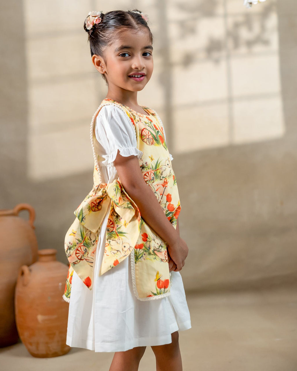 LAAD`S Girl in floral apron dress with a bunny and tulip print, layered over a white puff-sleeve dress. Features ruffled side panels and delicate lace trim. Soft, breathable fabric for comfort and charm.