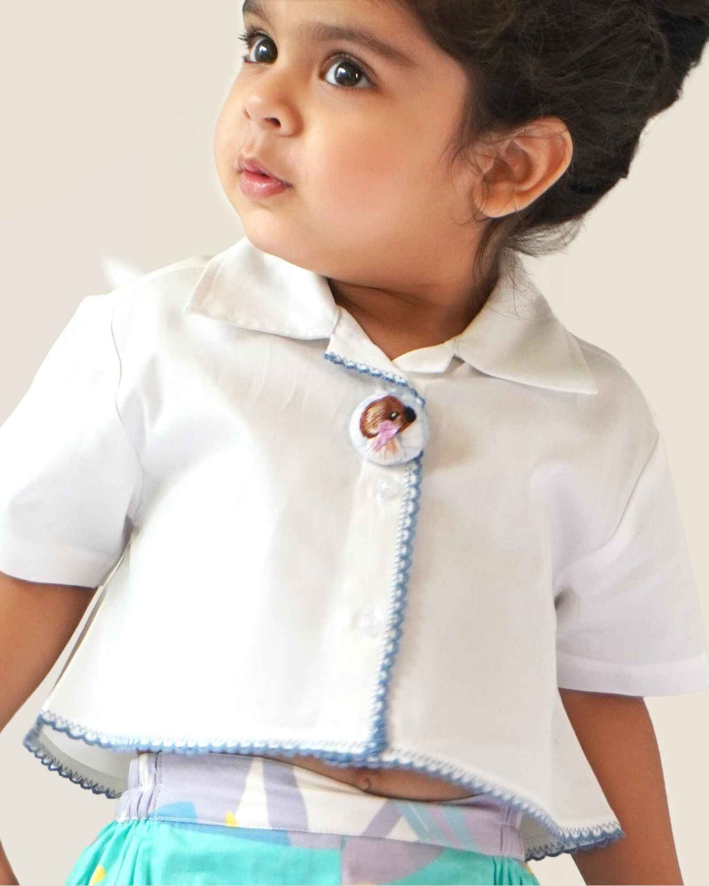 baby girl wearing  Laad`s gwhite cropped shirt with scalloped trim and hand-embroidered button on it along with pastel aqua bubble shots featuring a pleated abstract design.