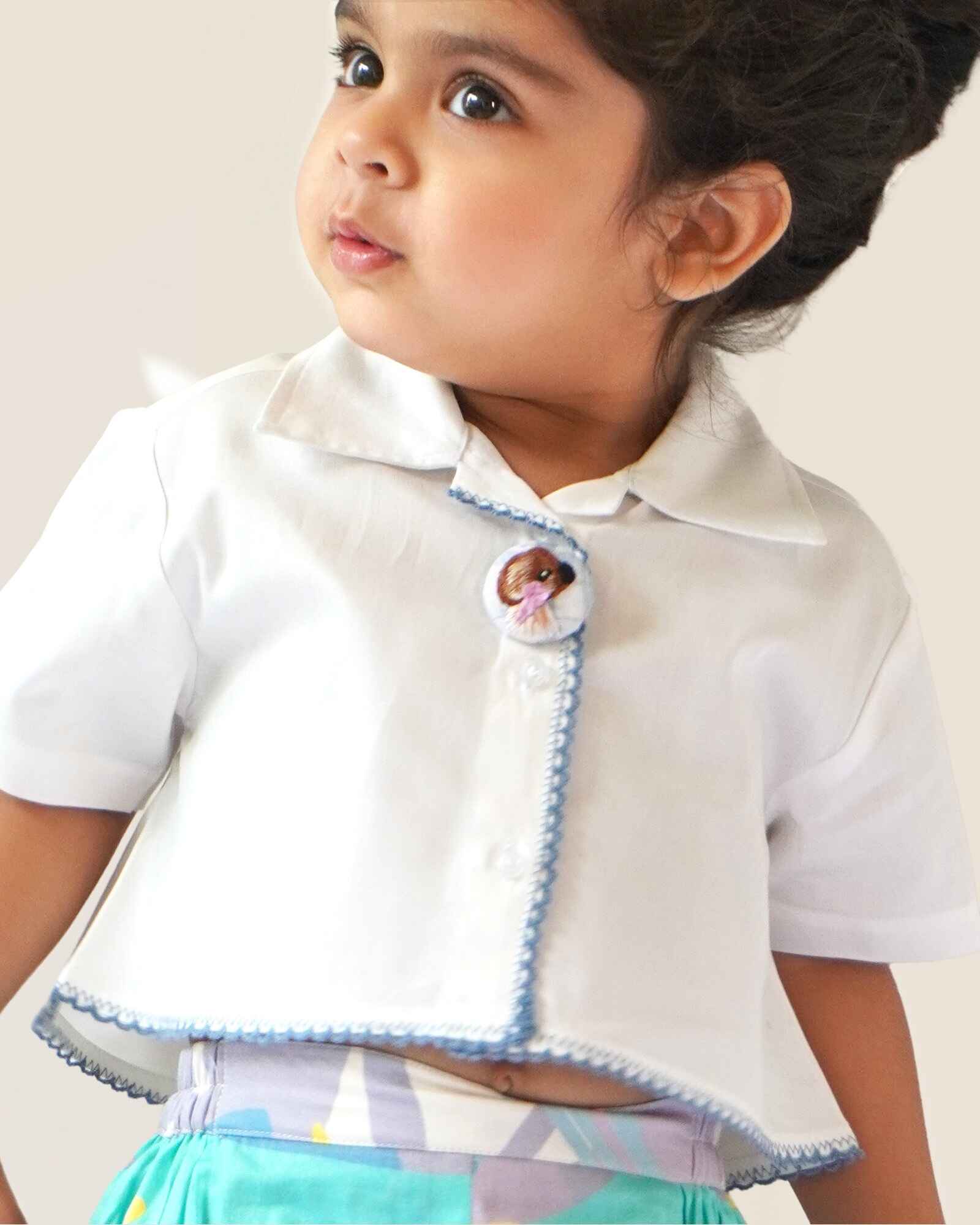 baby girl wearing  Laad`s gwhite cropped shirt with scalloped trim and hand-embroidered button on it along with pastel aqua bubble shots featuring a pleated abstract design.