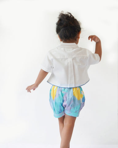 baby girl wearing  Laad`s gwhite cropped shirt with scalloped trim and hand-embroidered button on it along with pastel aqua bubble shots featuring a pleated abstract design.