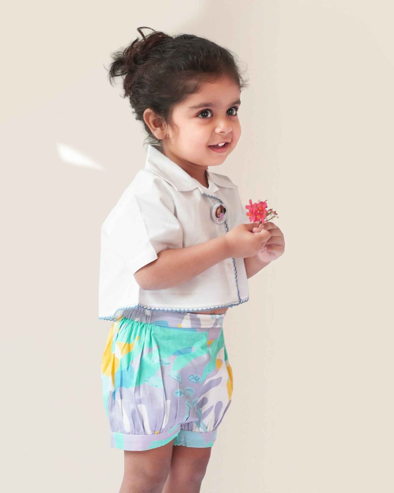baby girl wearing  Laad`s gwhite cropped shirt with scalloped trim and hand-embroidered button on it along with pastel aqua bubble shots featuring a pleated abstract design.