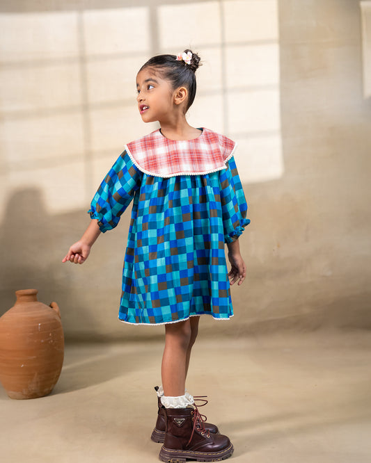 Girl wearing laad`s blue checkered girl blue bell dress with a red plaid Peter Pan collar and lace trim at the hem.