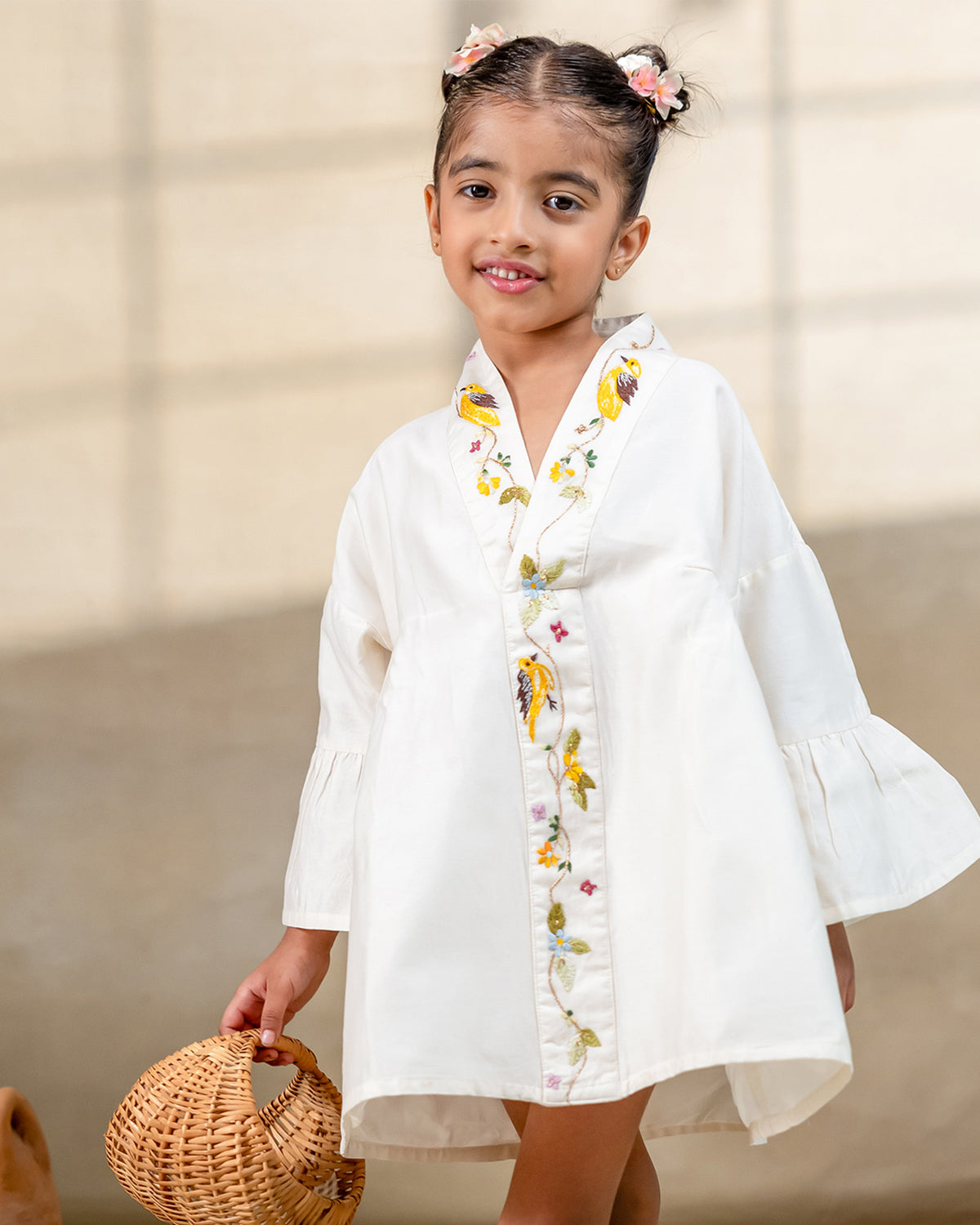 Little girl in a white hand-embroidered A-line dress with flared sleeves and colorful bird and floral detailing, holding a wicker basket.