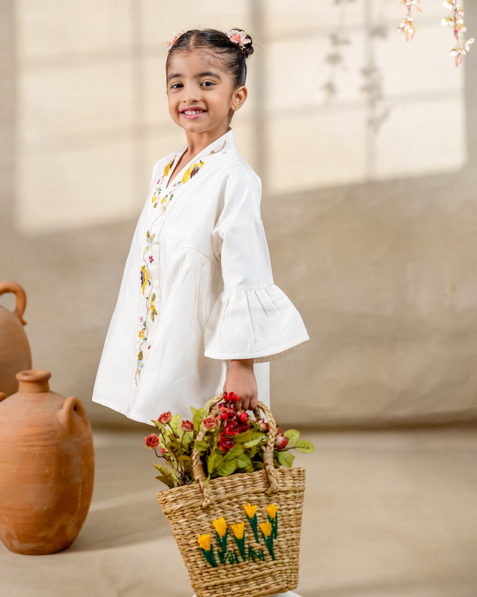 Little girl in a white hand-embroidered A-line dress with flared sleeves and colorful bird and floral detailing, holding a wicker basket.