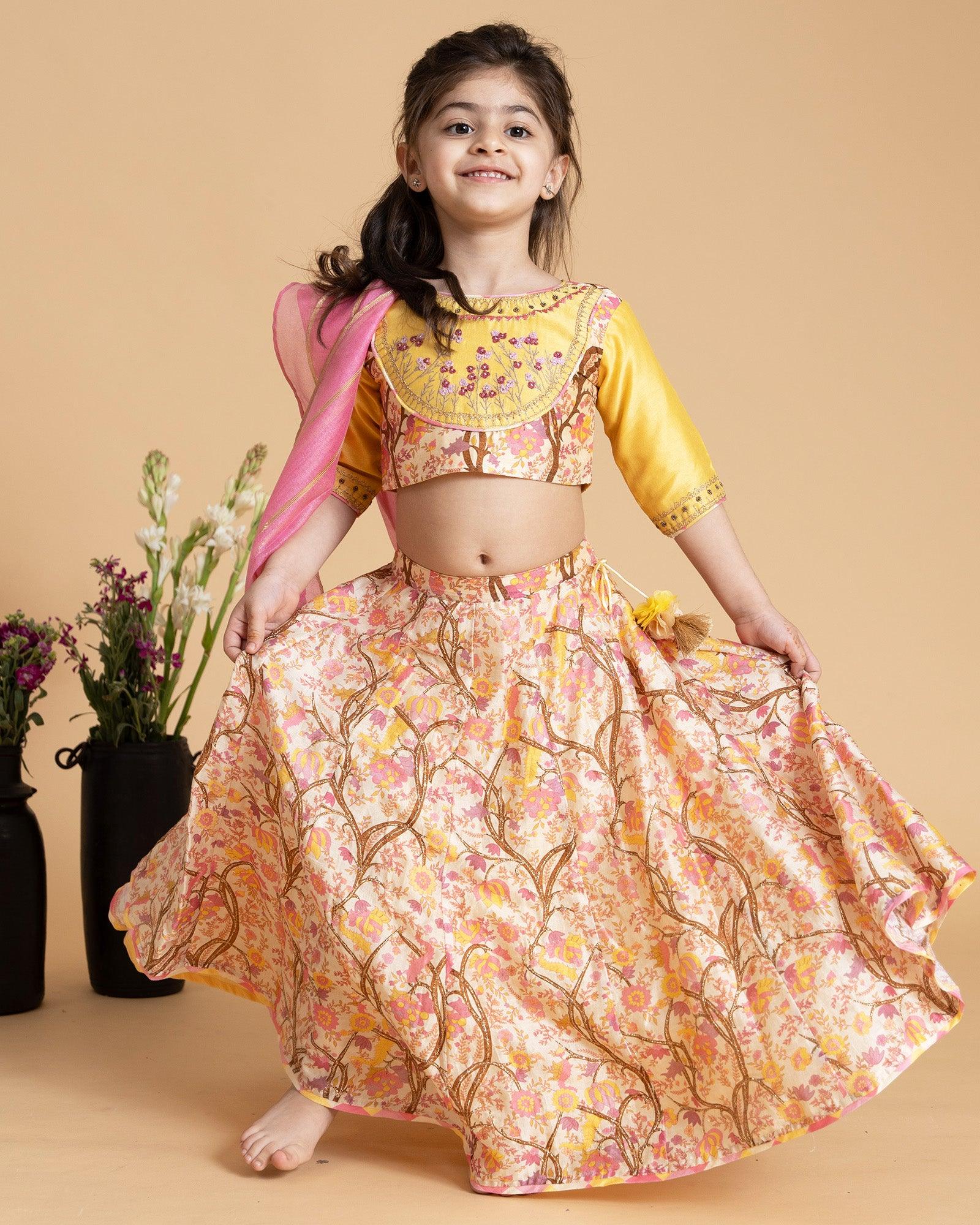 A girl in a floral embroidered blouse and skirt with badla work, tassel tie-up, and a pink lurex dupatta, exuding festive charm.
