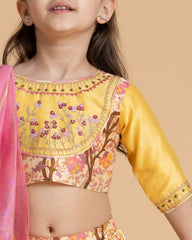 A girl in a floral embroidered blouse and skirt with badla work, tassel tie-up, and a pink lurex dupatta, exuding festive charm.