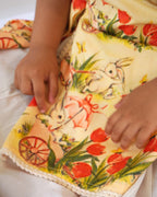 LAAD`S Girl in floral apron dress with a bunny and tulip print, layered over a white puff-sleeve dress. Features ruffled side panels and delicate lace trim. Soft, breathable fabric for comfort and charm.