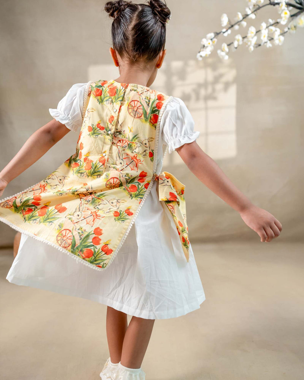 LAAD`S Girl in floral apron dress with a bunny and tulip print, layered over a white puff-sleeve dress. Features ruffled side panels and delicate lace trim. Soft, breathable fabric for comfort and charm.