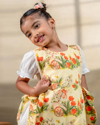 LAAD`S Girl in floral apron dress with a bunny and tulip print, layered over a white puff-sleeve dress. Features ruffled side panels and delicate lace trim. Soft, breathable fabric for comfort and charm.