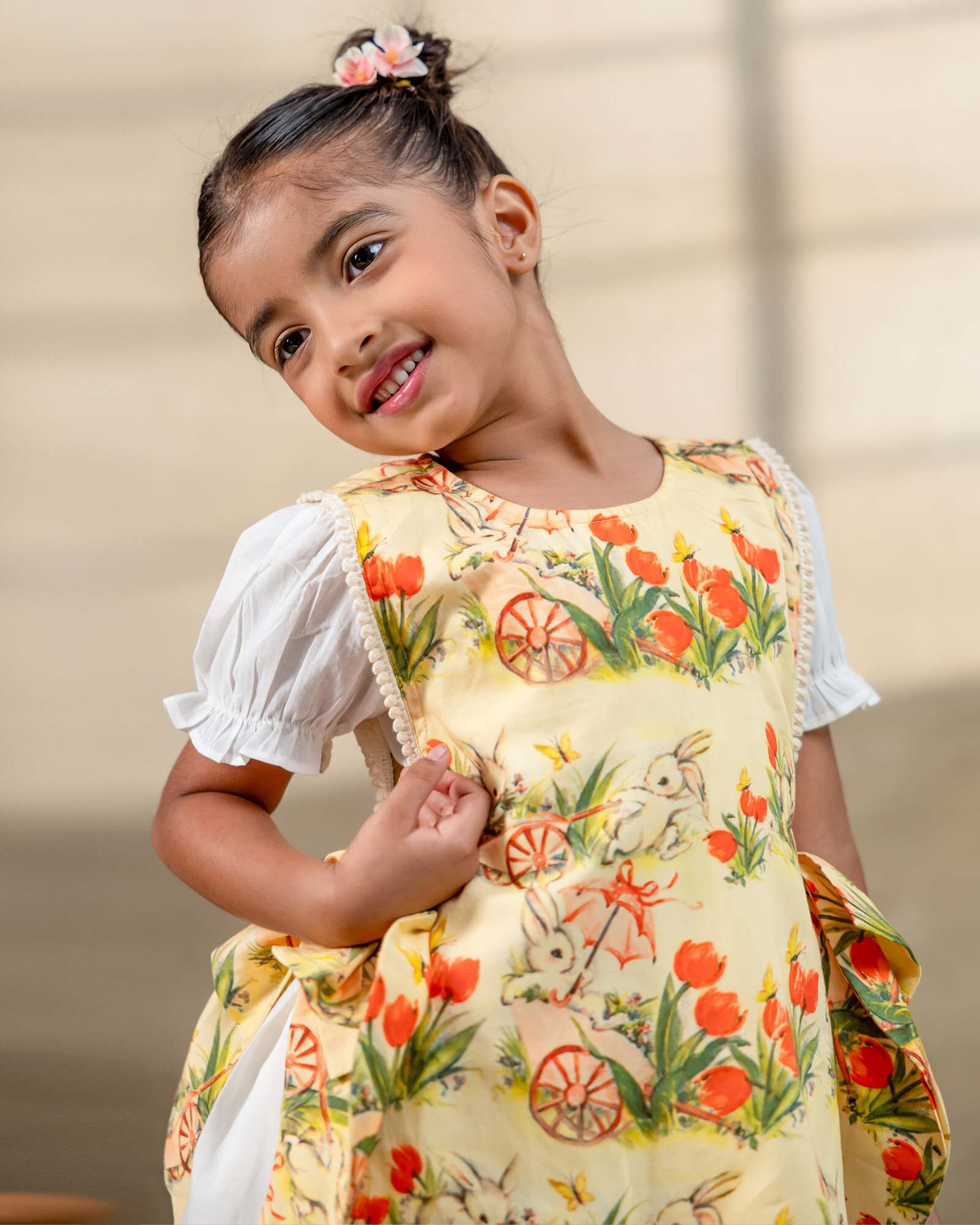 LAAD`S Girl in floral apron dress with a bunny and tulip print, layered over a white puff-sleeve dress. Features ruffled side panels and delicate lace trim. Soft, breathable fabric for comfort and charm.